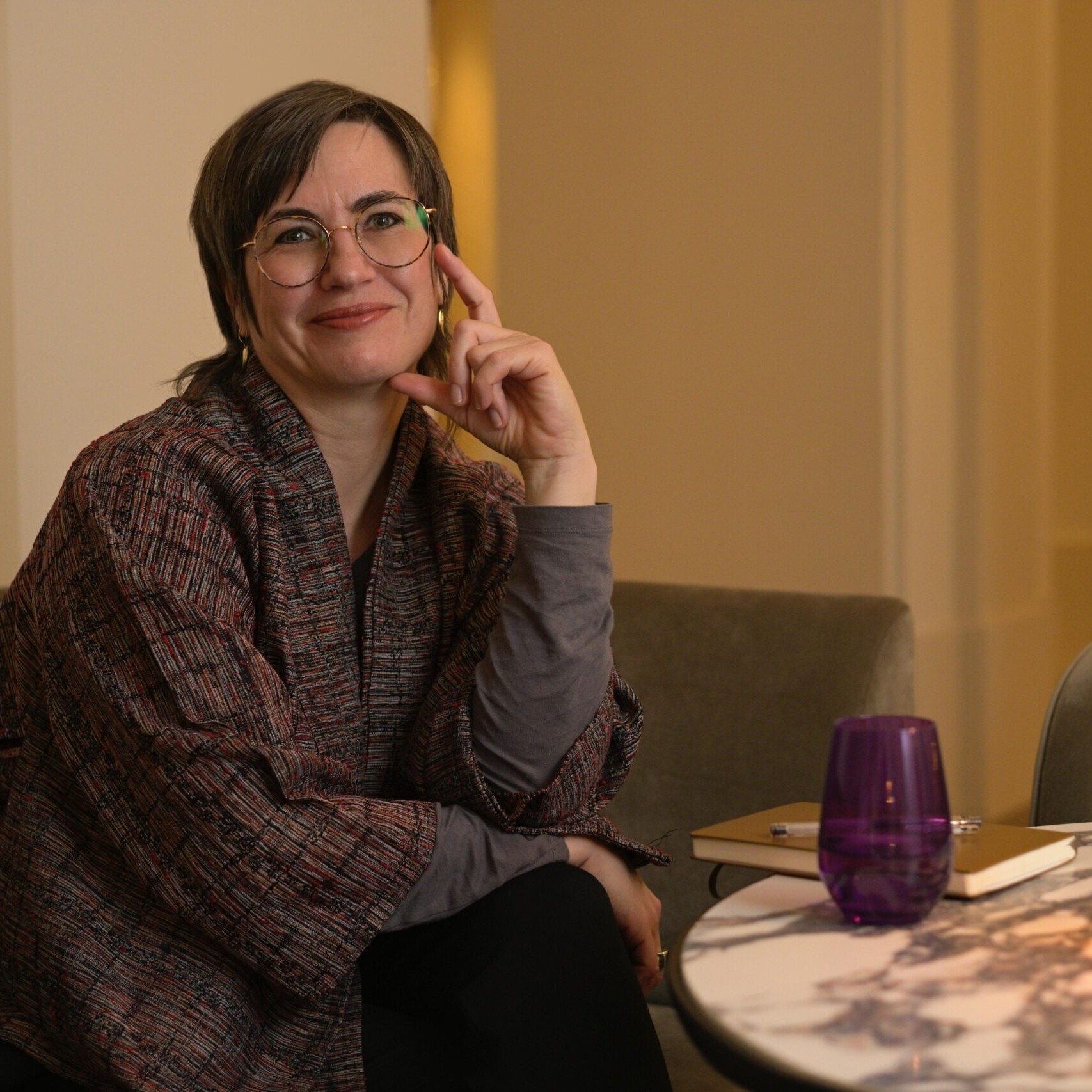 Die K&uuml;nstlerin Britta Jonas sitzt an einem runden kleinen Marmortisch auf dem eine kleine Lampe und ein lila Glas steht, ein Notizbuch mit Stift liegt daneben. Sie tr&auml;gt eine japanische handgewebte Jacke und schaut in die Camera.