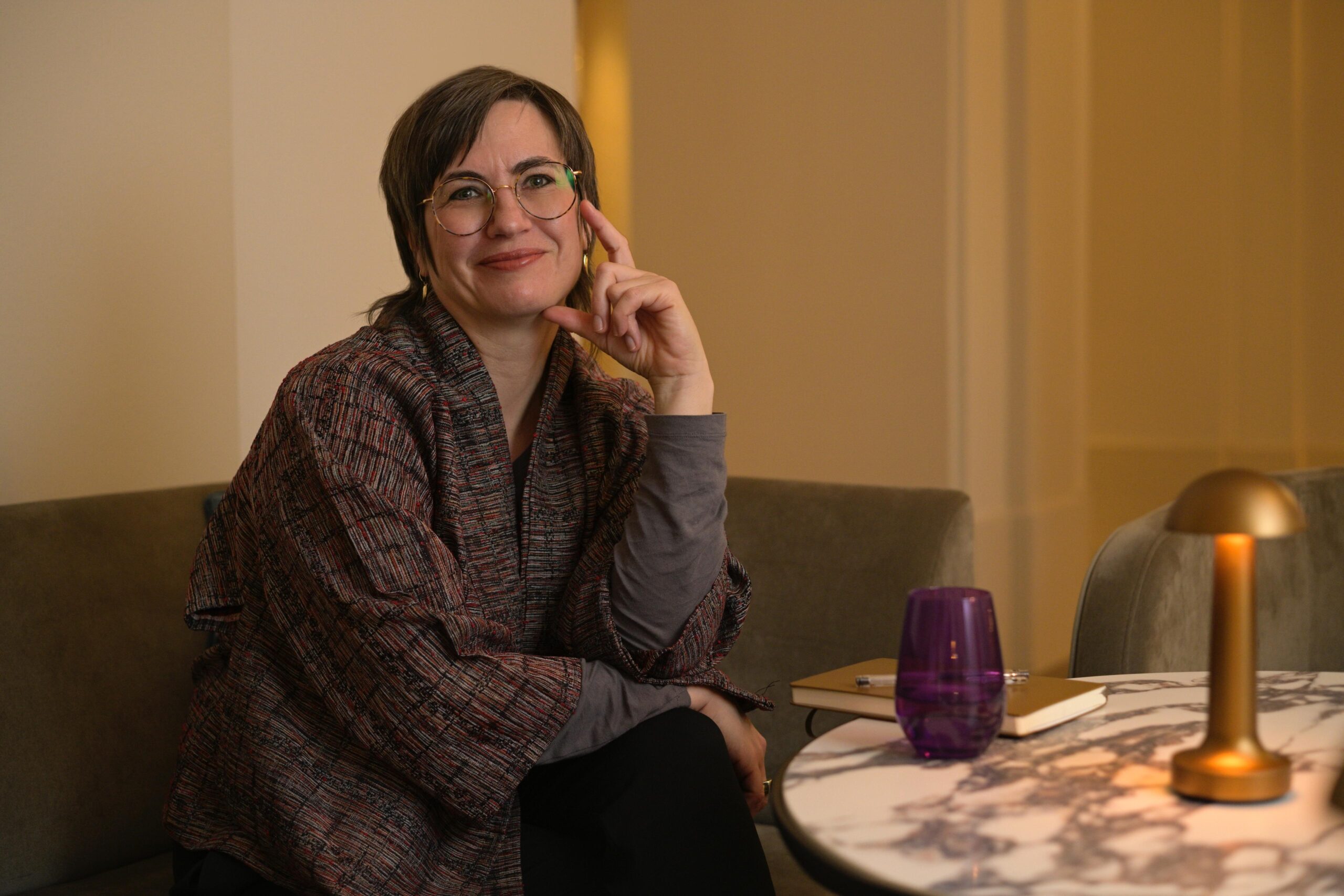 Die K&uuml;nstlerin Britta Jonas sitzt an einem runden kleinen Marmortisch auf dem eine kleine Lampe und ein lila Glas steht, ein Notizbuch mit Stift liegt daneben. Sie tr&auml;gt eine japanische handgewebte Jacke und schaut in die Camera.