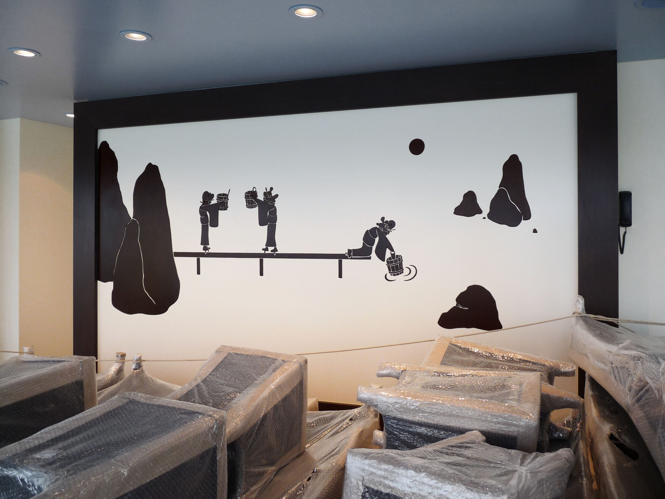 Wandgestaltung mit Landschaft und Wassertragenden und schöpfenden Geisha-Figuren. Davor sind in Folie eingepackte Stühle gelagert.